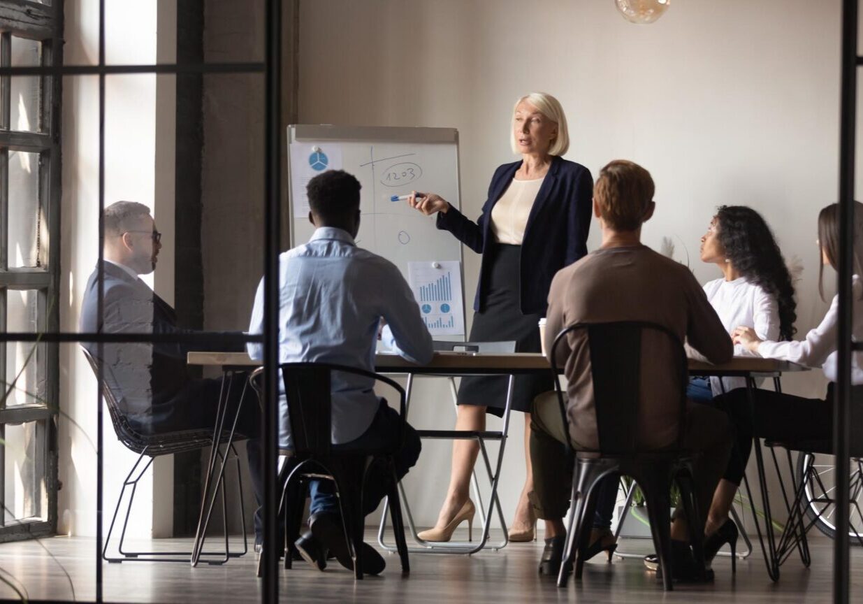 Mature older businesswoman mentor coach training speaker give flip chart business presentation at company meeting, confident female executive manager teaching team consult clients at office workshop
